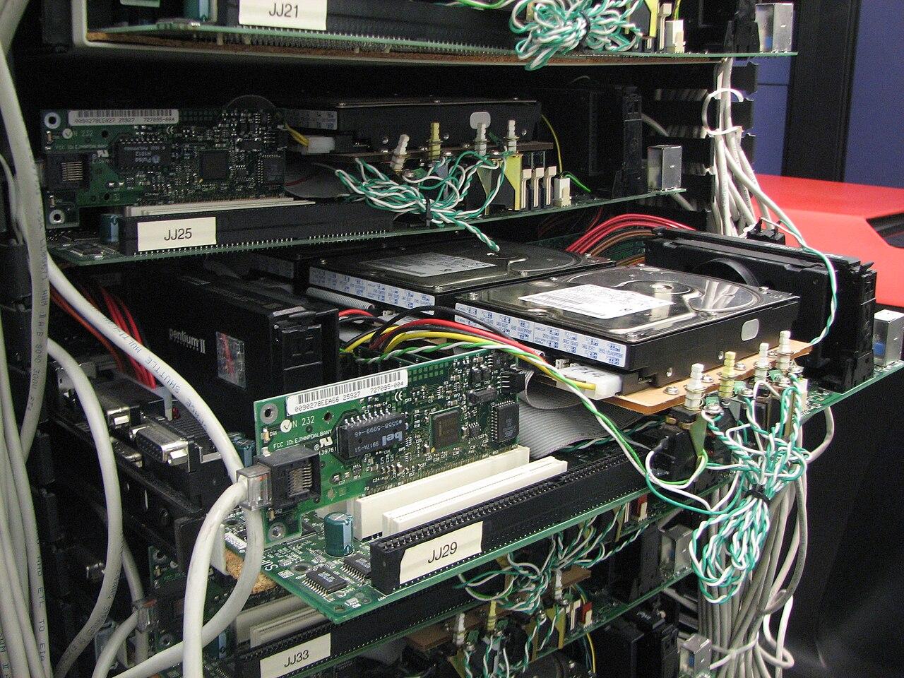 Google's first production server rack from 1998 — an open cabinet of stacked bare motherboards separated by corkboard trays — on display at the Computer History Museum.